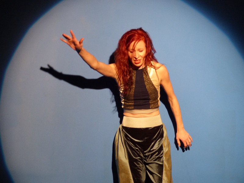 A woman in a silk tank top and flowy pants thrusts her arm out while standing in front of a blue wall. A spotlight illuminates her.